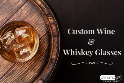 A custom promotional whiskey glass from Dynamic Gift sitting on a wooden barrel.